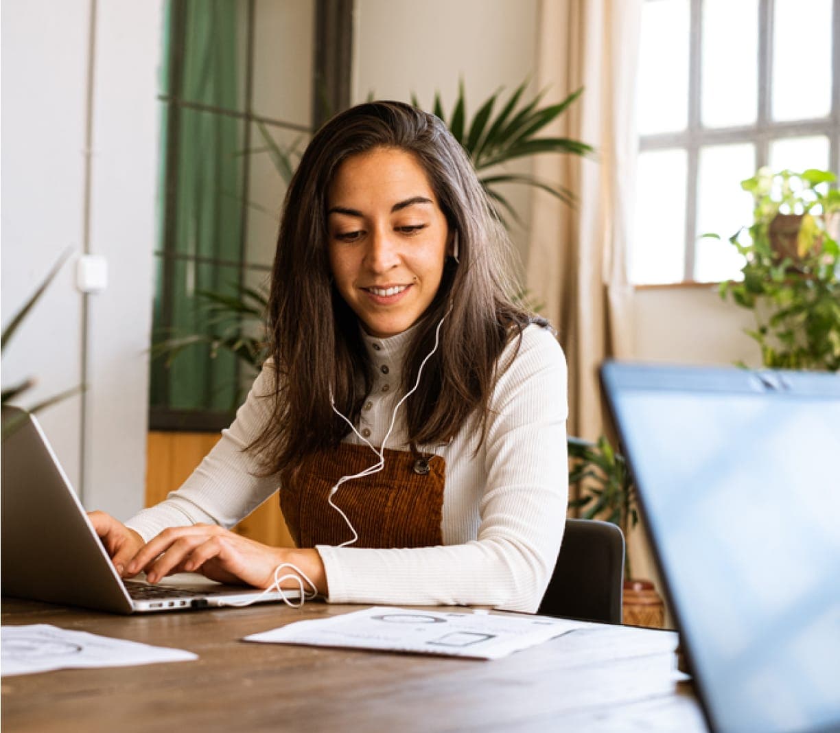 Smiling woman typing on computer wearing earphones looking at document while sitting at table