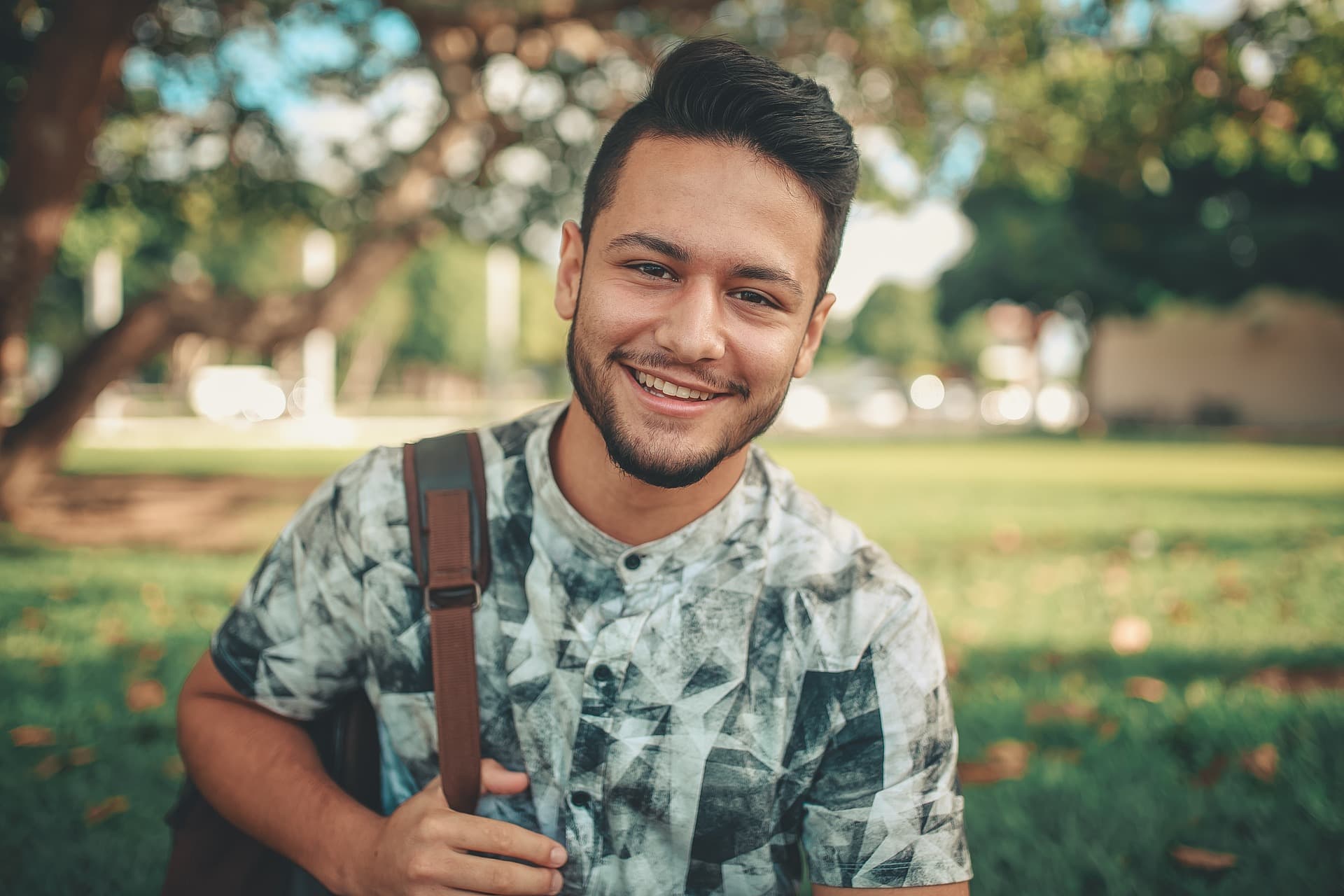 young man with backpack slung over one shoulder smiling outside on lawn in front of tree