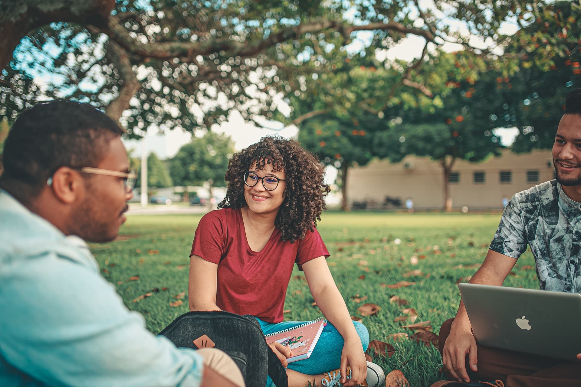 group of students sitting outside on grass under tree in circle smiling with one laptop computer