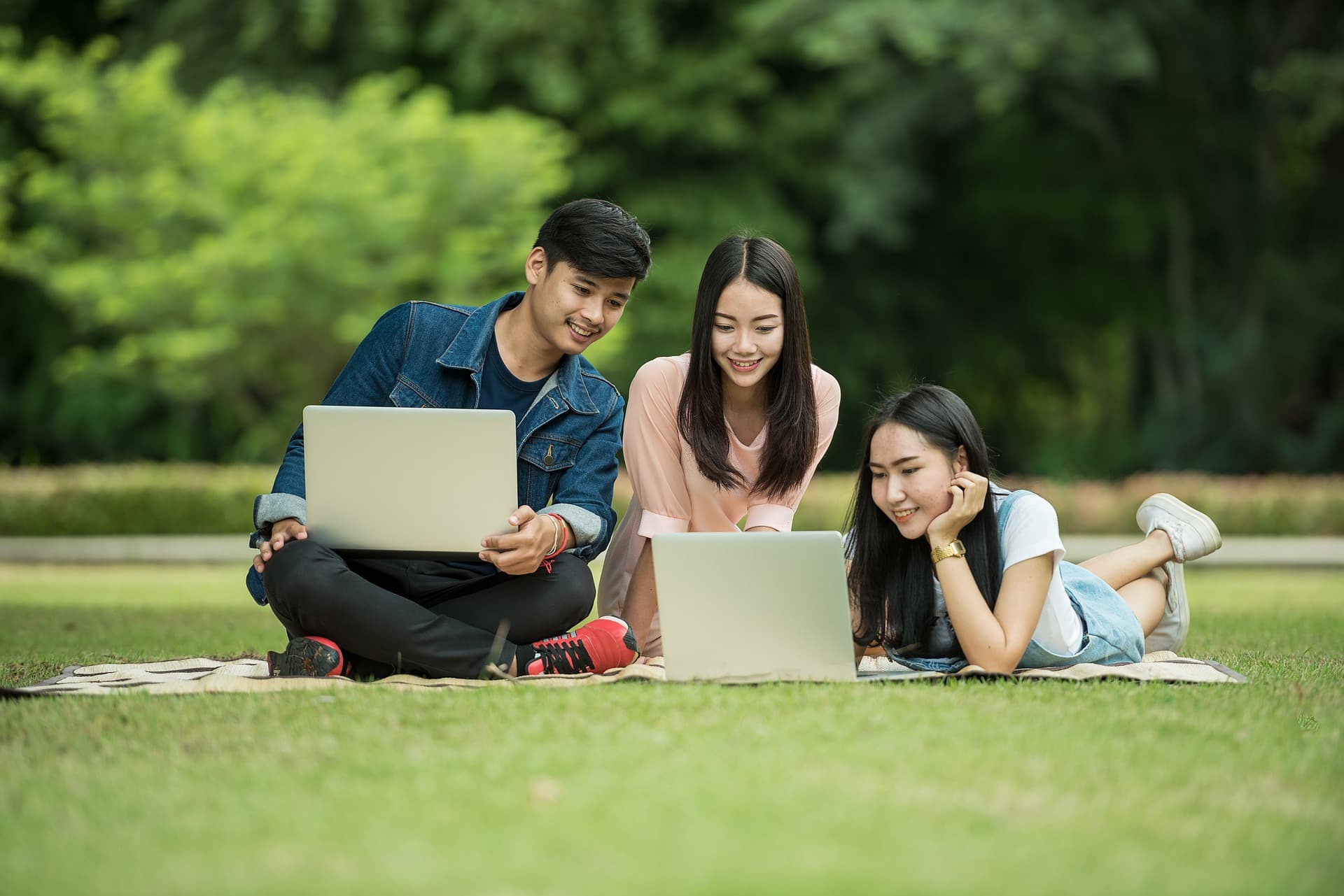 three students outside sitting on grass looking at one of two laptop computers