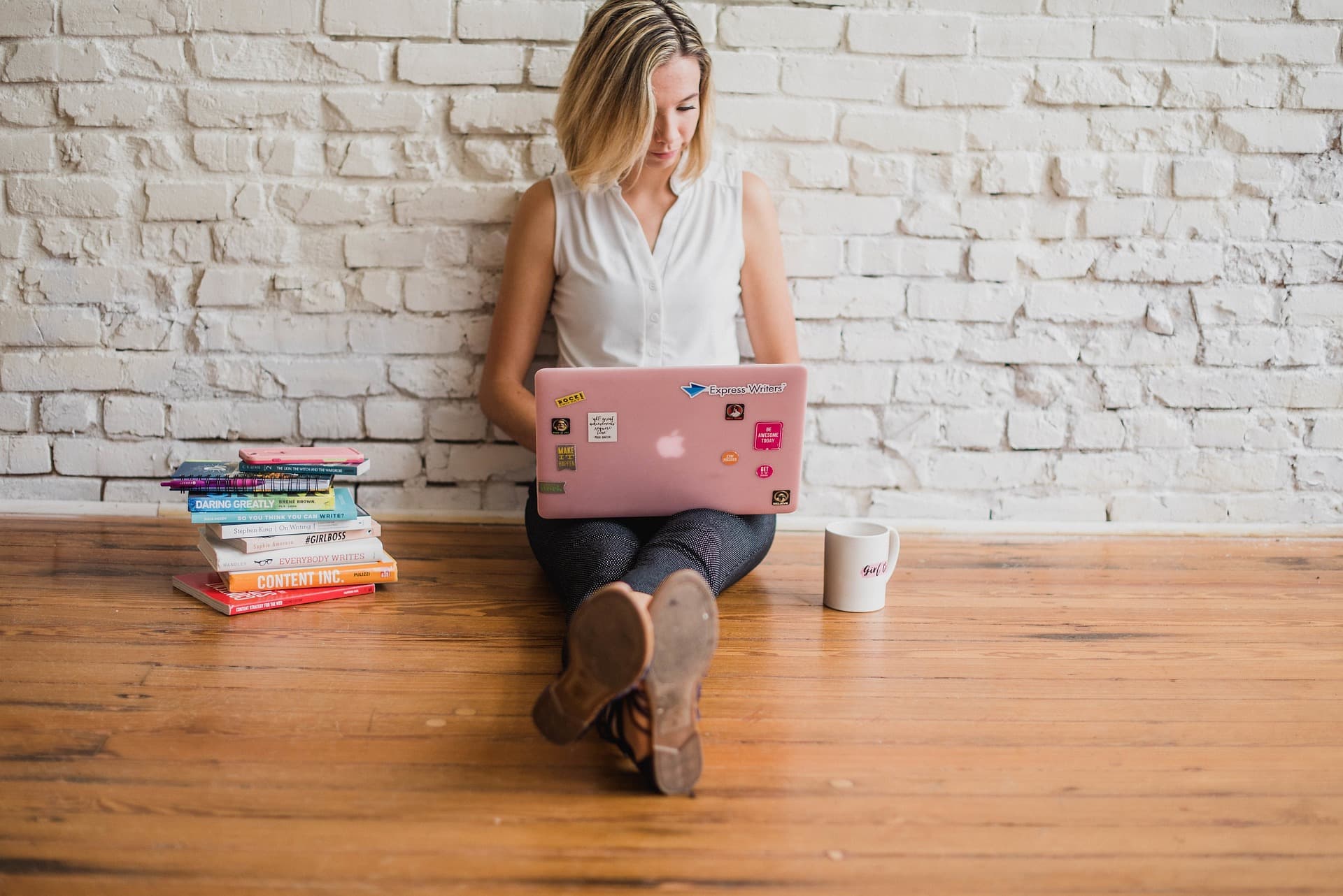 woman sitting on ground against wall typing on laptop beside pile of books