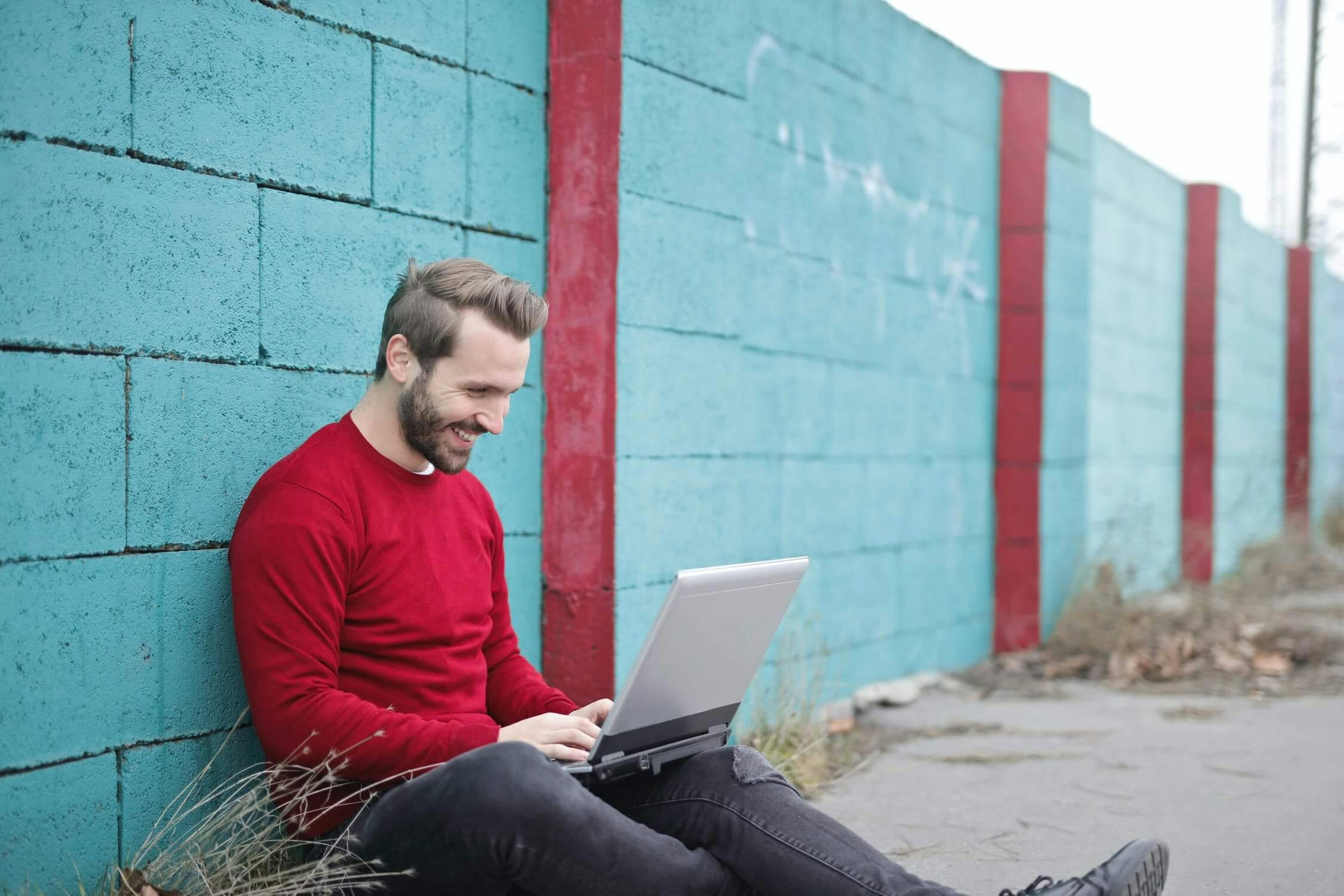 man sitting against cinderblock wall working on laptop