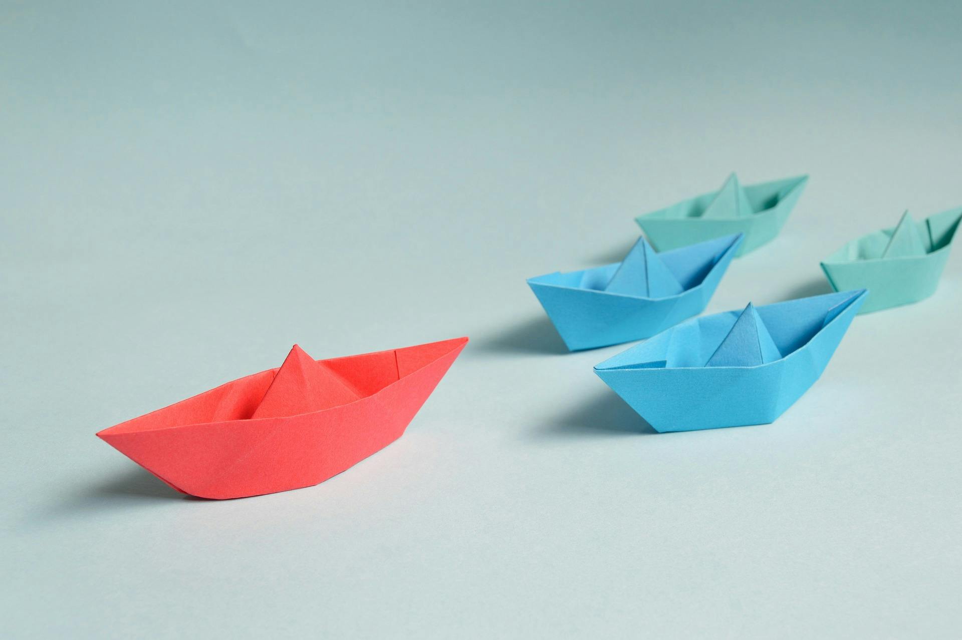 one red paper boat leading two blue paper boats leading two green paper boats