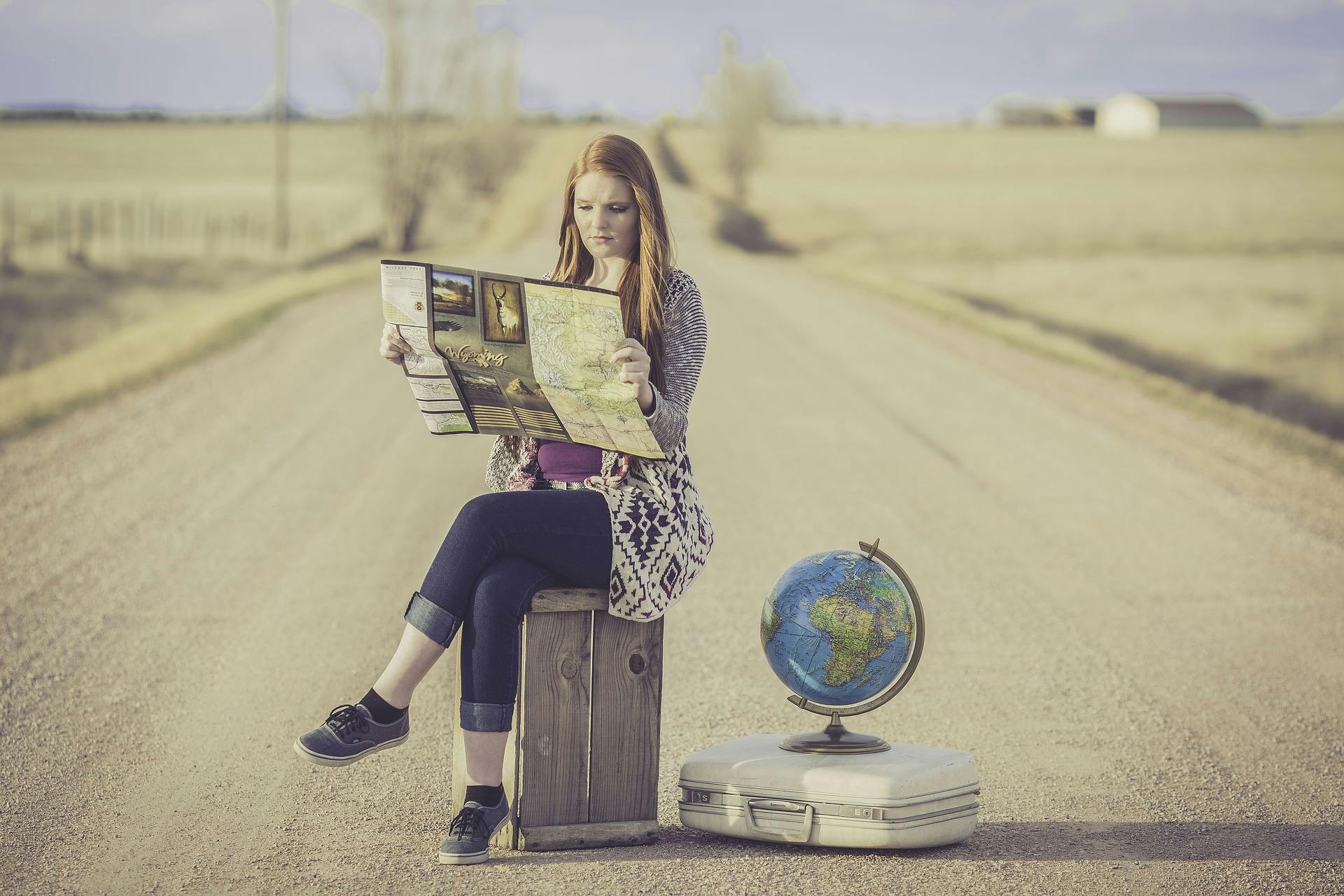 woman sitting on suitcase reading map on country road beside second suitcase with globe resting on top of it